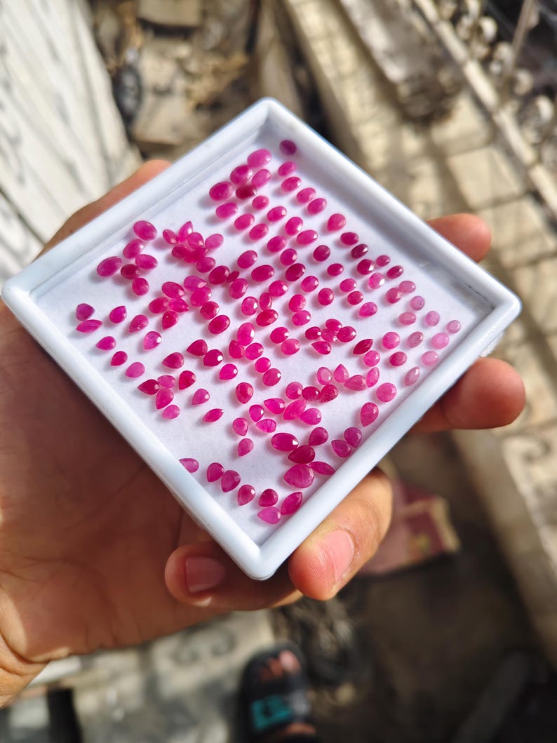 May include: A white square display tray containing a large collection of small, faceted, vibrant pink and red gemstones. The stones are arranged in a loose cluster on a white foam insert, featuring various shapes including oval, pear, and round cuts.