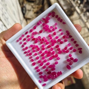 May include: A white rectangular display tray containing a large collection of small, loose, faceted rubies. The gemstones are vibrant pinkish-red in colour and feature various oval and pear shapes, arranged across the white foam insert.