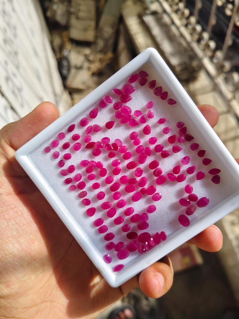 May include: A white rectangular display tray containing a large collection of small, loose, faceted rubies. The gemstones are vibrant pinkish-red in colour and feature various oval and pear shapes, arranged across the white foam insert.