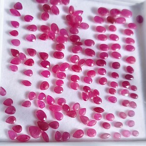 May include: A collection of numerous small, loose, faceted pinkish-red gemstones arranged on a white display tray. The stones vary in shape, including oval, pear, and round cuts, all exhibiting a vibrant, translucent, and polished appearance.