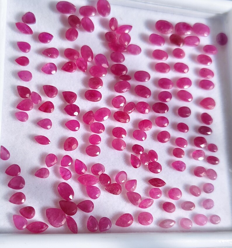 May include: A collection of numerous small, loose, faceted pinkish-red gemstones arranged on a white display tray. The stones vary in shape, including oval, pear, and round cuts, all exhibiting a vibrant, translucent, and polished appearance.