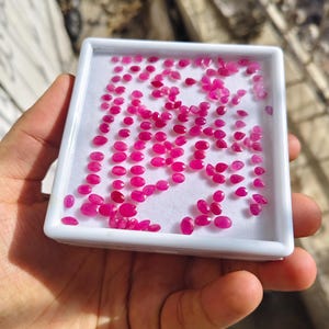 May include: A white rectangular display tray filled with numerous small, faceted, vibrant pinkish-red gemstones. The stones are arranged in a loose grid pattern on a white foam insert, showcasing various oval and pear shapes with reflective surfaces.