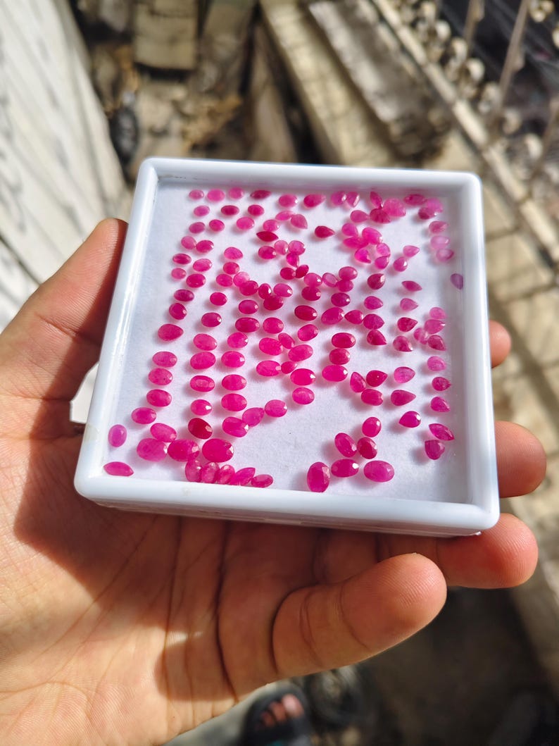 May include: A white rectangular display tray filled with numerous small, faceted, vibrant pinkish-red gemstones. The stones are arranged in a loose grid pattern on a white foam insert, showcasing various oval and pear shapes with reflective surfaces.