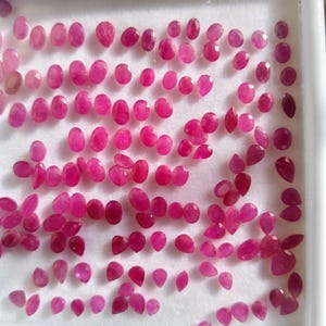 Pode incluir: Uma coleção de numerosos rubis pequenos, soltos e facetados em várias formas ovais e de pera. As pedras preciosas exibem uma cor vermelho-rosada vibrante e profunda, dispostas em grade numa bandeja de exibição de espuma branca.