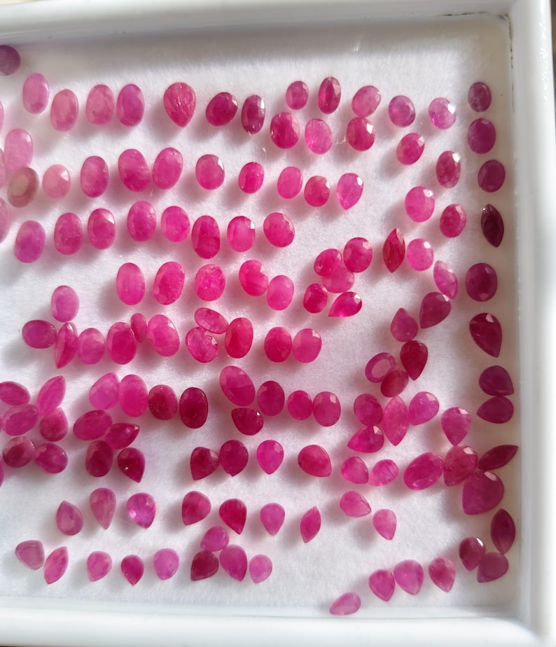 May include: A collection of numerous small, loose, faceted rubies in various oval and pear shapes. The gemstones exhibit a vibrant, deep pinkish-red colour and are arranged in a grid-like pattern on a white foam display tray with a white frame.