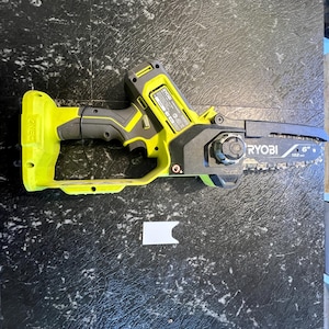 Puede incluir: Una motosierra inalámbrica amarilla y negra con una hoja de 15 cm. La marca "RYOBI" es visible. Esta herramienta manual está diseñada para cortar madera y otros materiales.
