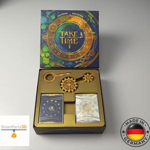 Könnte beinhalten: Ein Brettspielkarton mit dem Titel Take Time, dessen dunkelblauer Deckel eine goldene Uhr und Libellen zeigt. Die geöffnete Schachtel enthält einen goldenen Schlüssel, zwei zahnradförmige Spielsteine mit Augensymbolen und zwei Kartendecks.