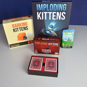 Może przedstawiać: Kolekcja pudełek z grą karcianą Exploding Kittens, w tym Imploding Kittens, Barking Kittens oraz mniejszy pakiet Streaking Kittens. Otwarte pudełko zawiera dwie talie czerwono-czarnych kart do gry z logo Exploding Kittens.
