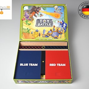 May include: A board game box titled Toy Battle with an open lid displaying a colorful illustration of a dinosaur, skeleton, and knight. The interior contains a wooden tray with circular tokens, and two large rectangular cards labeled Blue Team and Red Team.