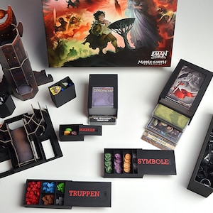 Puede incluir: Un juego de mesa con varias cajas de almacenamiento negras, tarjeteros y una torre. La caja del juego presenta ilustraciones con figuras y el texto "ZMAN Middle-earth Collection". El juego incluye piezas y cartas.