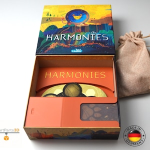 Peut inclure: Boîte de jeu ouverte avec le mot "HARMONIES" en lettres dorées. La boîte présente une illustration colorée d'un lion et d'un paysage. Un petit sac en toile de jute est à côté de la boîte. Fabriqué en Allemagne.