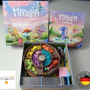 Könnte beinhalten: Das Brettspiel Mycelia und die Prisma-Erweiterungsbox sind zusammen mit einer offenen Spielschachtel abgebildet, die einen braunen Kunststoffeinsatz mit gelben, orangen, grünen, violetten und blauen Spielsteinen sowie Kartenstapeln enthält.