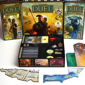 Op de afbeelding: Drie bordspeldozen getiteld "7 Wonders Duel" met de uitbreidingen Agora, Pantheon en Duel. De dozen bevatten illustraties van oude beschavingen. Spelcomponenten zijn zichtbaar in een zwarte organizer.