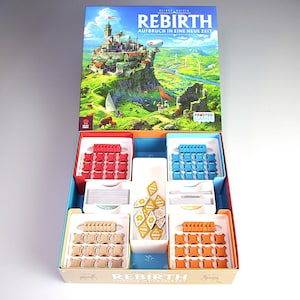 Op de afbeelding: Een bordspeldoos getiteld Rebirth Aufbruch in eine neue Zeit met een open deksel met een kleurrijke landschapsillustratie. De binnenkant bevat georganiseerde plastic bakjes met rode, blauwe, beige en oranje speelstukken en kaarten.