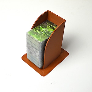 Può includere: Un porta carte marrone con base quadrata e lati curvi, che contiene una pila di carte. Le carte hanno un'immagine verde e bianca. Il supporto è realizzato in materiale solido e poggia su una superficie bianca.