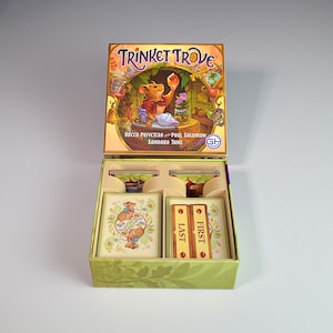 Peut inclure: Une boîte de jeu de société intitulée Trinket Trove avec une illustration de souris tenant une gemme orange brillante. La boîte ouverte contient des jeux de cartes organisés, avec deux jetons rectangulaires marqués First et Last.