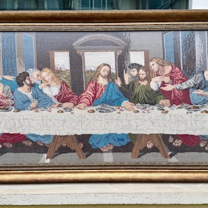 Può includere: Un arazzo incorniciato raffigurante l'Ultima Cena. L'opera presenta figure in abiti sedute attorno a una lunga tavola coperta da una tovaglia bianca. La cornice è dorata e lo sfondo mostra dettagli architettonici.