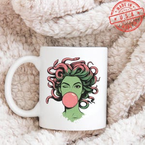May include: White ceramic mug featuring a graphic of Medusa winking and blowing a bubblegum bubble. The illustration has green skin, pink hair, and snakes. The mug has a white handle and the words "MADE IN U.S.A." in a red stamp.