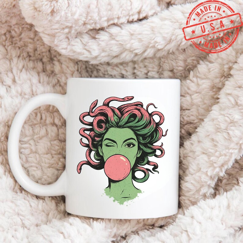May include: White ceramic mug featuring a graphic of Medusa winking and blowing a bubblegum bubble. The illustration has green skin, pink hair, and snakes. The mug has a white handle and the words "MADE IN U.S.A." in a red stamp.