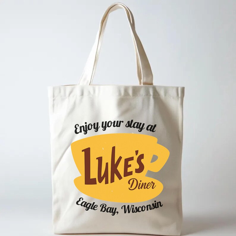 May include: Cream-colored tote bag with a yellow coffee cup design and the words "Luke's Diner" in brown. The text "Enjoy your stay at Eagle Bay, Wisconsin" is also printed on the bag. The bag has two cream-colored handles.