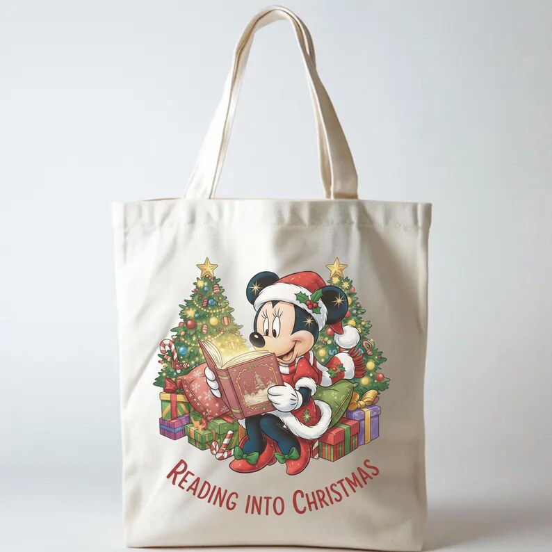 K&ouml;nnte beinhalten: Beige Tragetasche mit einer weihnachtlichen Illustration von Minnie Mouse, die ein Buch liest. Sie tr&auml;gt einen Weihnachtsmannhut und ein rotes Outfit, umgeben von Geschenken und Weihnachtsb&auml;umen. Der Text "Reading into Christmas" ist auf der Tasche aufgedruckt.
