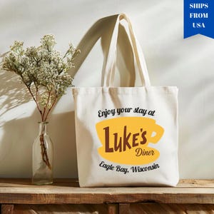 May include: Cream-colored tote bag with the text "Enjoy your stay at Luke's Diner" in black script. A yellow coffee cup graphic with the words "Luke's Diner" in brown is centered on the bag. The text "Eagle Bay, Wisconsin" is below.