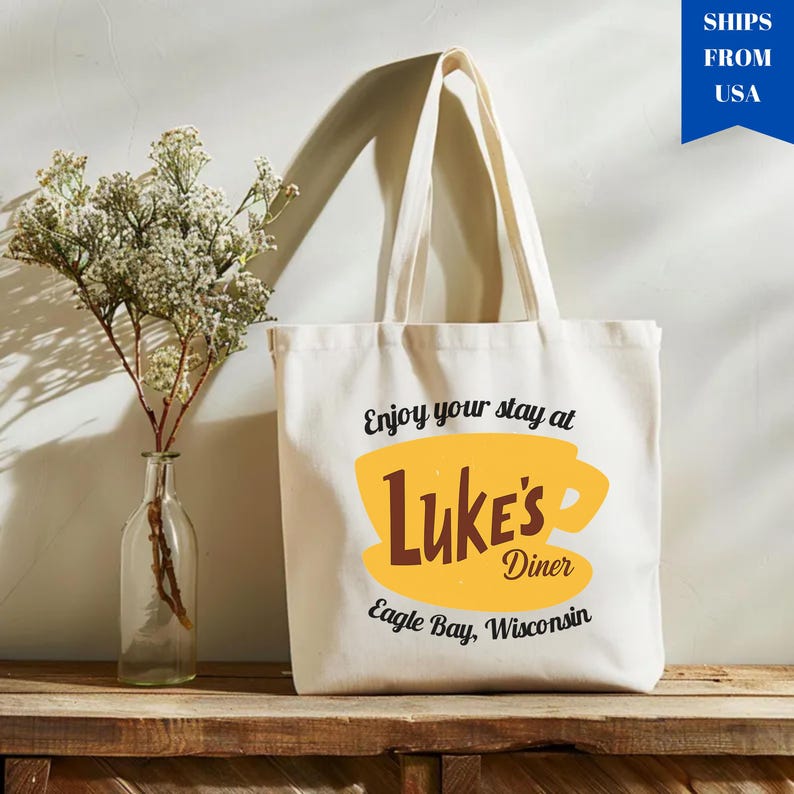May include: Cream-colored tote bag with the text "Enjoy your stay at Luke's Diner" in black script. A yellow coffee cup graphic with the words "Luke's Diner" in brown is centered on the bag. The text "Eagle Bay, Wisconsin" is below.