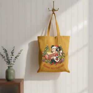 K&ouml;nnte beinhalten: Senfgelbe Tragetasche mit einer weihnachtlichen Illustration von Mickey Mouse, die ein Buch liest. Mickey tr&auml;gt einen Weihnachtsmannhut und ist von Geschenken und Weihnachtsb&auml;umen umgeben. Der Text "READING INTO CHRISTMAS" ist darunter gedruckt.