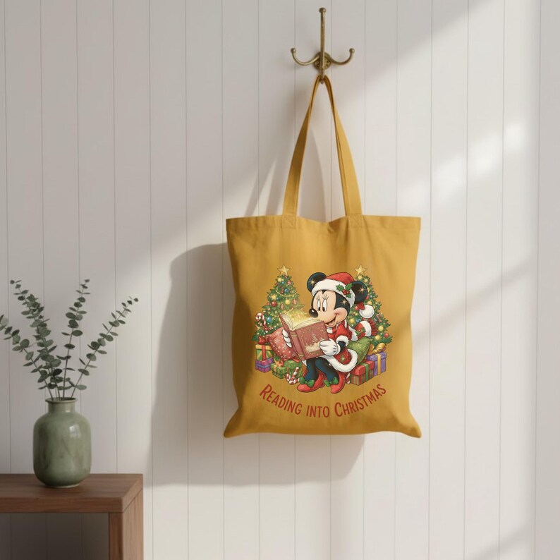 K&ouml;nnte beinhalten: Senfgelbe Tragetasche mit einer weihnachtlichen Illustration von Mickey Mouse, die ein Buch liest. Mickey tr&auml;gt einen Weihnachtsmannhut und ist von Geschenken und Weihnachtsb&auml;umen umgeben. Der Text "READING INTO CHRISTMAS" ist darunter gedruckt.