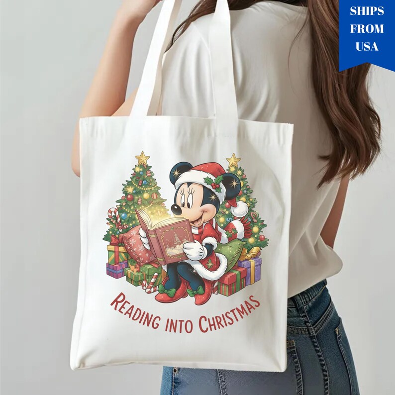 K&ouml;nnte beinhalten: Wei&szlig;e Tragetasche mit einer farbenfrohen Illustration von Minnie Mouse, die vor Weihnachtsb&auml;umen und Geschenken ein Buch liest. Minnie Mouse tr&auml;gt einen Weihnachtsmannhut und ein rotes Outfit. Der Text "Reading into Christmas" ist unter dem Bild gedruckt.
