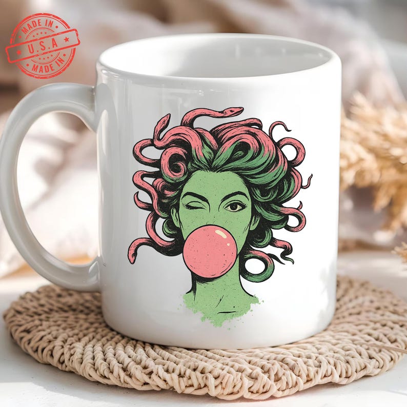 May include: White ceramic mug featuring a Medusa illustration with green skin, pink and green hair, and a pink bubblegum bubble. The mug has a large handle and a "MADE IN U.S.A." stamp.