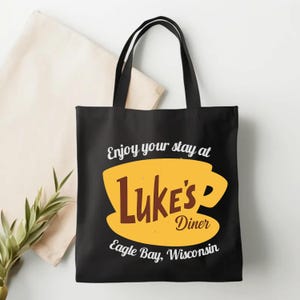 May include: Black tote bag featuring a yellow coffee cup design with the words "Luke's Diner" in brown and white text. The text "Enjoy your stay at" and "Eagle Bay, Wisconsin" are also displayed.