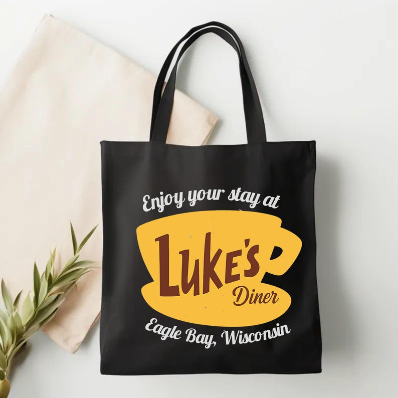 May include: Black tote bag featuring a yellow coffee cup design with the words "Luke's Diner" in brown and white text. The text "Enjoy your stay at" and "Eagle Bay, Wisconsin" are also displayed.