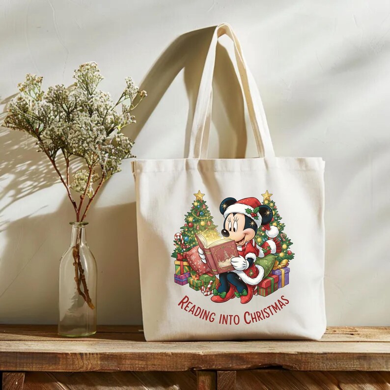 K&ouml;nnte beinhalten: Cremefarbene Tragetasche mit einem weihnachtlichen Motiv von Mickey Mouse, der ein Buch liest. Mickey tr&auml;gt einen Weihnachtsmannhut und -anzug, umgeben von Weihnachtsb&auml;umen und Geschenken. Der Text "Reading into Christmas" ist darunter gedruckt.