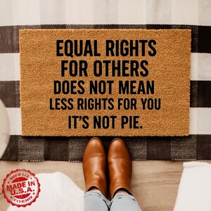 May include: A rectangular doormat with the text "EQUAL RIGHTS FOR OTHERS DOES NOT MEAN LESS RIGHTS FOR YOU IT'S NOT PIE." in black. The mat is brown and sits on a black and white striped rug. Brown boots are in front of the mat.