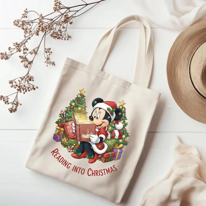 K&ouml;nnte beinhalten: Beige Tragetasche mit einer weihnachtlichen Illustration einer Comic-Maus, die ein Buch liest. Die Maus tr&auml;gt einen Weihnachtsmannhut und ein rotes Outfit, umgeben von Weihnachtsb&auml;umen und Geschenken. Der Text "READING INTO CHRISTMAS" ist auf der Tasche aufgedruckt.