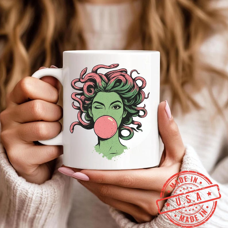 May include: White ceramic mug featuring a graphic of a green-skinned woman with pink snakes for hair, winking and blowing a pink bubble. The mug has a white handle and is held by a person. A red "Made in U.S.A." stamp is visible.