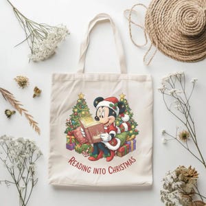 K&ouml;nnte beinhalten: Cremefarbene Tragetasche mit einer festlichen Illustration von Minnie Mouse mit Weihnachtsm&uuml;tze, die vor Weihnachtsb&auml;umen und Geschenken ein Buch liest. Der Text "Reading into Christmas" ist unter dem Bild aufgedruckt. Die Tasche hat zwei Henkel.