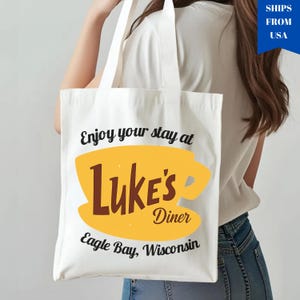 May include: White tote bag featuring the text "Enjoy your stay at Luke's Diner" with a yellow coffee cup design and the words "Luke's Diner" in brown. The text "Eagle Bay, Wisconsin" is also included.