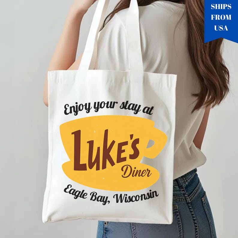May include: White tote bag featuring the text "Enjoy your stay at Luke's Diner" with a yellow coffee cup design and the words "Luke's Diner" in brown. The text "Eagle Bay, Wisconsin" is also included.