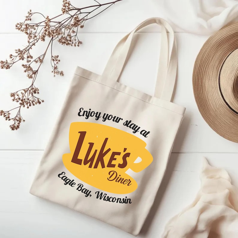 May include: Cream-colored tote bag with the text "Enjoy your stay at Luke's Diner Eagle Bay, Wisconsin" in black and brown. The design includes a yellow coffee cup graphic. A straw hat and dried flowers are in the background.