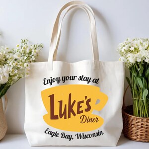 May include: Cream-colored tote bag with the text "Enjoy your stay at Luke's Diner" in black, with a yellow coffee cup graphic and the words "Luke's Diner" in brown. The text "Eagle Bay, Wisconsin" is below.