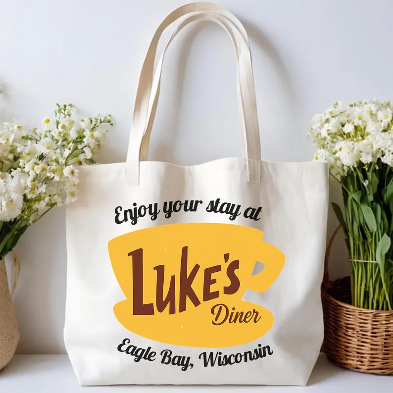 May include: Cream-colored tote bag with the text "Enjoy your stay at Luke's Diner" in black, with a yellow coffee cup graphic and the words "Luke's Diner" in brown. The text "Eagle Bay, Wisconsin" is below.