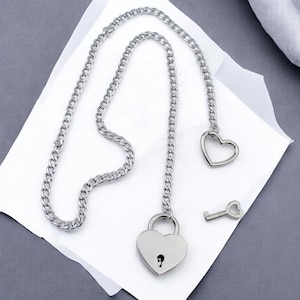 Könnte beinhalten: Silberfarbene Halskette mit herzförmigem Vorhängeschloss-Anhänger, herzförmigem Charm und Schlüssel-Charm. Die Halskette hat ein Panzerketten-Design. Die Artikel werden auf einer weißen Oberfläche vor grauem Hintergrund präsentiert.