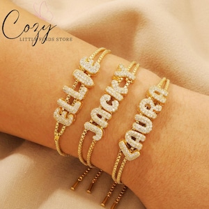 Puede incluir: Tres pulseras doradas con los nombres "ELLA", "JACK" y "LAURA" escritos con cristales transparentes brillantes. Las pulseras están en una muñeca, sobre un fondo beige neutro. Las pulseras tienen un diseño ajustable.