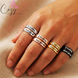 May include: Five rings are shown in silver, rose gold, gold, and black. Each ring is engraved with the words "I fucking love you" and a heart symbol. The rings are a romantic and edgy jewelry item.
