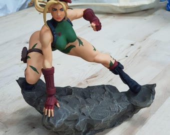 May include: A detailed figurine of a female character in a dynamic pose, wearing a green bodysuit with leaf-like accents, a red beret, and matching gloves. The figure is posed on a textured, gray rock base.