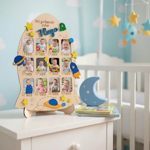 Puede incluir: Marco de fotos de madera con forma de cohete con espacios para 12 fotos de bebé, con meses. El marco tiene un diseño de planeta azul y amarillo, estrellas y el nombre "Hugo". Una luna azul y libros están sobre la mesa.
