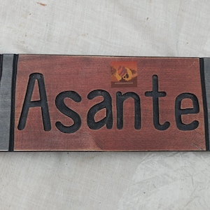 May include: A rectangular wooden sign with the word "Asante" carved in large, bold letters. The sign is brown with black lettering and decorative edges. The sign has a rustic, handcrafted appearance.