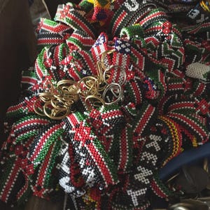 May include: A selection of beaded items in red, black, green, and white. The beads are arranged in geometric patterns. Gold and silver wire jewellery is also present. The items appear to be handmade and are likely decorative or wearable.